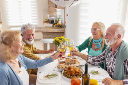Foto de Dos parejas mayores divirtiéndose mientras celebran Acción de Gracias juntas en casa durante la cena tradicional, levantando copas de vino, haciendo un brindis - Imagen libre de derechos