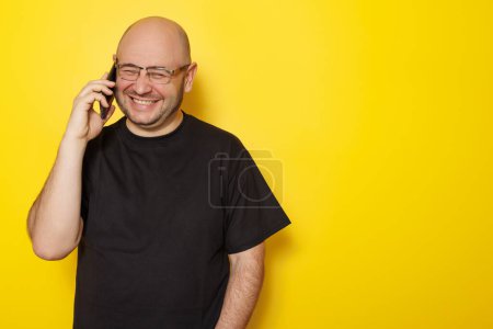 Téléchargez les photos : Mi-trentaine chauve homme en surpoids portant un t-shirt noir et des lunettes ayant une conversation téléphonique en utilisant un téléphone intelligent isolé sur fond jaune avec un espace de copie - en image libre de droit