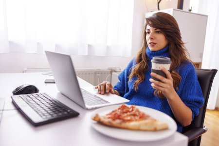 Téléchargez les photos : Programmeuse assise à son bureau à la maison, prenant une pause tout en travaillant à distance de la maison en buvant du café et en mangeant de la pizza - en image libre de droit
