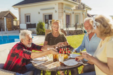 Photo for Group of senior friends having lunch in the backyard by the swimming pool, gathered around the table, eating, drinking and relaxing outdoors on a sunny summer day - Royalty Free Image
