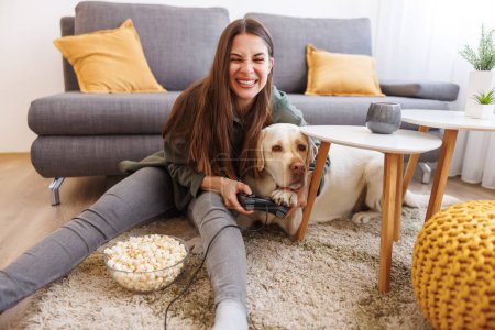 Téléchargez les photos : Jeune femme s'amusant passer du temps libre à la maison avec son animal de compagnie, beau chien du Labrador, jouer à des jeux vidéo et manger du maïs soufflé - en image libre de droit
