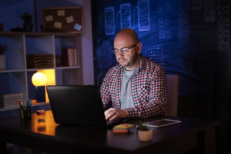 Téléchargez les photos : Développeur Web assis à son bureau tout en travaillant tard dans un bureau à domicile, en utilisant un ordinateur portable - en image libre de droit