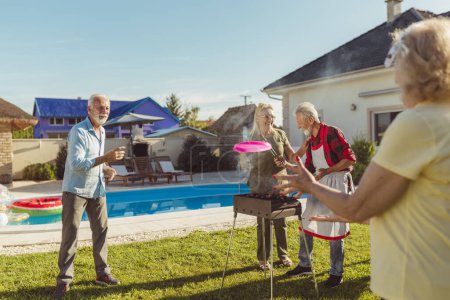 Téléchargez les photos : Groupe de personnes âgées actives gaies ayant une fête barbecue arrière-cour, griller de la viande et des légumes et avoir du plaisir à jouer lancer et attraper - en image libre de droit