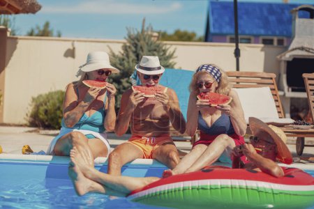 Téléchargez les photos : Groupe de personnes âgées gaies assis au bord d'une piscine bronzer, manger de la pastèque et s'amuser à l'extérieur par une chaude journée d'été - en image libre de droit