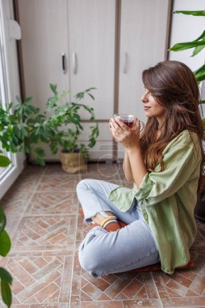 Téléchargez les photos : Belle jeune femme relaxante à la maison assise sur son balcon en buvant du thé chaud - en image libre de droit