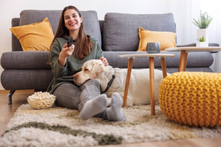Photo for Beautiful young woman relaxing at home, sitting on the living room floor watching TV and eating popcorn while her dog is lying next to her with head in her lap - Royalty Free Image