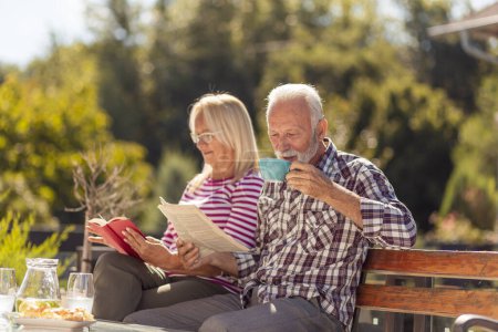 Photo for Happy senior couple enjoying their time together having an outdoor breakfast in the backyard of their home, man reading newspaper while woman is reading a book - Royalty Free Image