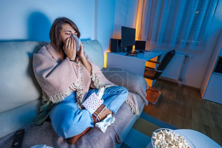 Téléchargez les photos : Vue en angle élevé d'une femme malade assise sur un canapé recouvert d'une couverture tard dans la nuit, ayant la grippe et la fièvre, se mouchant avec des mouchoirs en papier tout en regardant la télévision - en image libre de droit