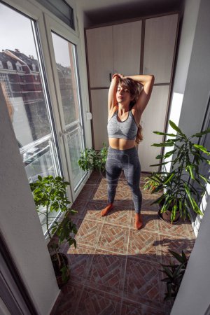 Téléchargez les photos : Jeune femme en vêtements de sport étirement tout en faisant de l'entraînement à la maison sur son balcon - en image libre de droit