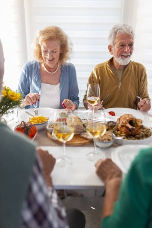 Foto de Grupo de amigos mayores divirtiéndose mientras se reúnen alrededor de la mesa para la cena tradicional de Acción de Gracias, comiendo, bebiendo y riendo, disfrutando de su tiempo juntos - Imagen libre de derechos