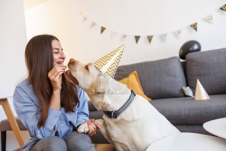 Foto de Hermosa joven que se divierte haciendo una fiesta de cumpleaños para su perro, usando sombreros de fiesta y dándole golosinas - Imagen libre de derechos