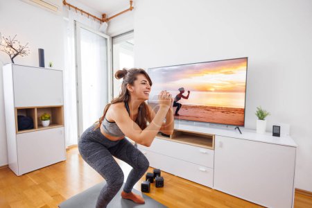 Photo for Active young woman doing squats while working out at home, following exercises shown by instructor on TV - Royalty Free Image