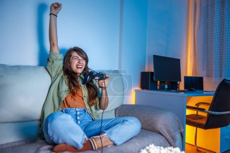 Téléchargez les photos : Belle jeune femme s'amuser à la maison tard dans la nuit à jouer à des jeux vidéo, célébrer après avoir gagné le jeu - en image libre de droit