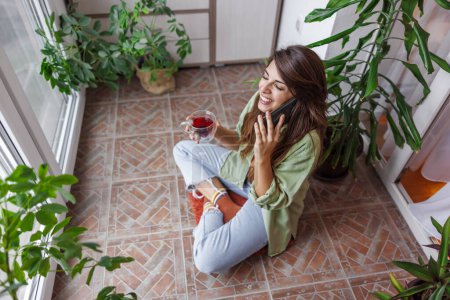Téléchargez les photos : Belle jeune femme jouissant de loisirs à la maison, ayant une conversation téléphonique avec un ami et buvant du thé tout en se relaxant sur son balcon - en image libre de droit