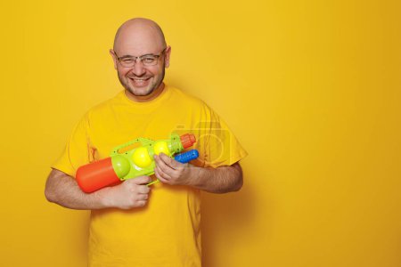 Foto de El hombre se divierte jugando con pistola de agua mientras se prepara para el viaje de vacaciones de verano en el fondo de color amarillo con espacio de copia - Imagen libre de derechos