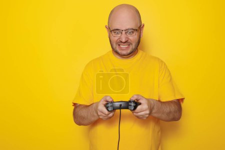 Téléchargez les photos : Joyeux milieu de la trentaine homme s'amusant à jouer à des jeux vidéo sur fond de couleur jaune avec espace de copie - en image libre de droit