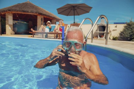 Foto de Grupo de amigos mayores relajándose y tomando el sol junto a la piscina, disfrutando de las vacaciones de verano; anciano poniéndose una máscara de snorkel, divirtiéndose haciendo snorkel en la piscina - Imagen libre de derechos