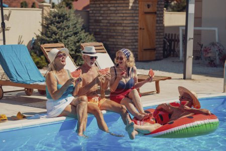 Photo for Group of cheerful senior people sitting at the edge of a swimming pool eating watermelon, sunbathing and having fun outdoors on a hot summer day - Royalty Free Image