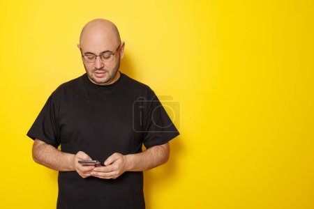 Photo for Mid 30s bald overweight man wearing black T-shirt and glasses typing text message using smart phone isolated on yellow colored background with copy space - Royalty Free Image