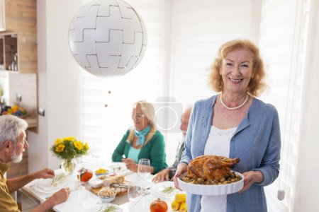 Photo for Senior friends gathered around table, celebrating Thanksgiving, having dinner together at home, hostess welcoming guests and serving roasted chicken - Royalty Free Image