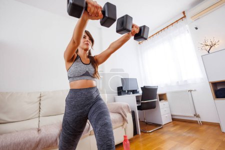 Téléchargez les photos : Jeune femme active en vêtements de sport faisant des exercices de bras en utilisant des poids à la maison - en image libre de droit