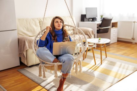 Téléchargez les photos : Femme relaxante à la maison, assise dans une chaise suspendue dans le salon, utilisant un ordinateur portable et un téléphone intelligent - en image libre de droit