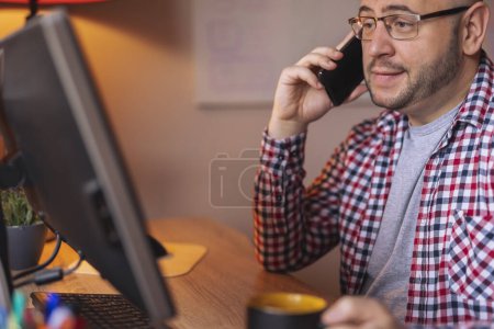 Téléchargez les photos : Homme freelance assis à son bureau avoir une conversation téléphonique et boire du café tout en travaillant au bureau à la maison - en image libre de droit