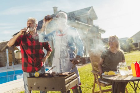 Photo for Two senior men drinking beer while grilling meat at backyard barbecue with their wives sitting at the table in the background, drinking coffee and relaxing - Royalty Free Image
