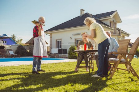Photo for Elderly couple having friends over for an outdoor lunch in the backyard by the swimming pool, serving food and drinks and having fun spending sunny summer day together - Royalty Free Image