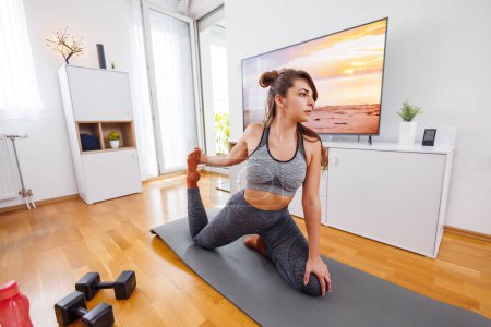 Foto de Mujer en forma activa en ropa deportiva que se extiende en la esterilla de yoga mientras hace ejercicio en casa - Imagen libre de derechos
