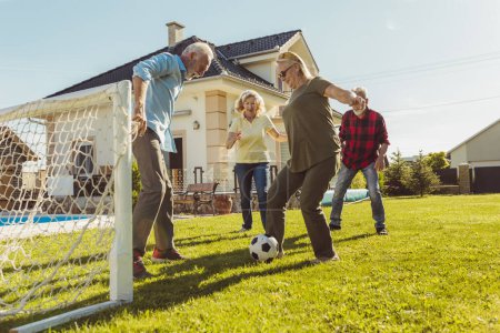 Foto de Grupo de personas mayores activas que se divierten jugando al fútbol en el césped en el patio trasero, disfrutando de un soleado día de verano al aire libre, corriendo tras una pelota - Imagen libre de derechos