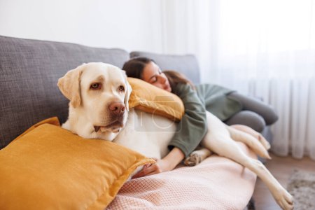 Foto de Mujer relajándose en casa tomando una siesta por la tarde acostada en el sofá durmiendo y abrazando a su perro mascota - Imagen libre de derechos