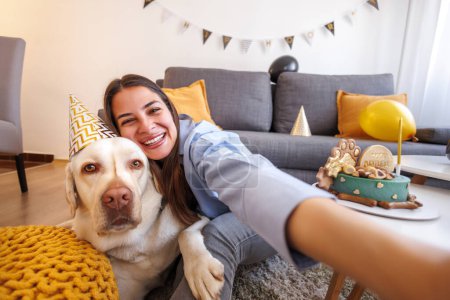 Foto de Hermosa joven alegre divirtiéndose tomando selfies mientras celebra el cumpleaños de su perro labrador mascota en casa - Imagen libre de derechos