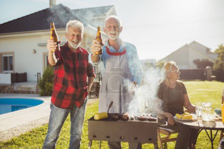 Téléchargez les photos : Deux hommes âgés font un toast avec des bouteilles de bière tout en grillant de la viande sur le barbecue arrière-cour avec leurs épouses assis à la table en arrière-plan, boire du café et se détendre - en image libre de droit