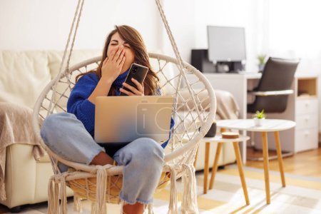 Téléchargez les photos : Jeune femme assise dans une chaise suspendue tout en travaillant à distance de la maison, bâillant et utilisant un téléphone intelligent, fatigué et endormi - en image libre de droit