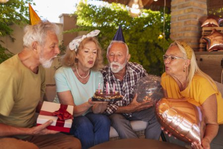 Foto de Grupo de alegres amigos mayores divirtiéndose en una fiesta de cumpleaños, usando sombreros de fiesta, sosteniendo globos y soplando velas en el pastel de cumpleaños - Imagen libre de derechos