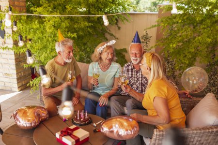 Foto de Grupo de amigos mayores alegres activos que usan sombreros de fiesta que se divierten celebrando cumpleaños, haciendo un brindis y bebiendo vino - Imagen libre de derechos