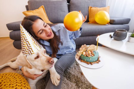 Téléchargez les photos : Joyeux jeune femme s'amuser à souffler des ballons tout en organisant une fête d'anniversaire pour son chien à la maison - en image libre de droit