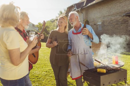 Téléchargez les photos : Groupe d'amis aînés joyeux faisant la fête barbecue arrière-cour, réunis autour du gril, jouant de la guitare, chantant, grillant de la viande et s'amusant - en image libre de droit