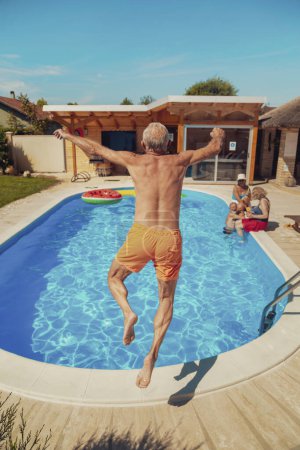 Téléchargez les photos : Groupe d'amis âgés se détendre et bronzer au bord de la piscine par une chaude journée d'été, un homme qui s'amuse à sauter dans la piscine pendant ses vacances - en image libre de droit