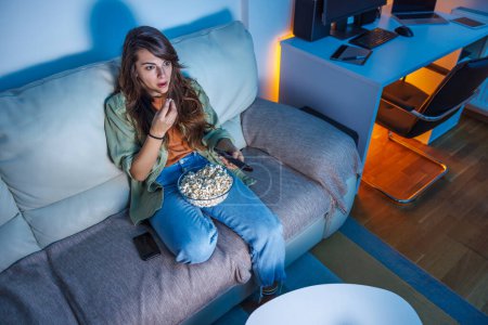 Téléchargez les photos : Belle jeune femme s'amusant à regarder la télévision et manger du pop-corn à la maison la nuit - en image libre de droit