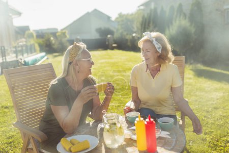 Foto de Dos ancianas sentadas en una mesa, almorzando al aire libre, relajándose y divirtiéndose en la fiesta de barbacoa del patio trasero - Imagen libre de derechos