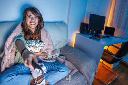 Téléchargez les photos : Belle jeune femme assise sur le canapé du salon recouvert d'une couverture relaxante à la maison la nuit en s'amusant à regarder la télévision et à manger du pop-corn - en image libre de droit
