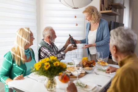 Foto de Dos parejas mayores divirtiéndose mientras celebran el Día de Acción de Gracias juntas en casa durante la cena tradicional, sirviendo vino y haciendo un brindis - Imagen libre de derechos