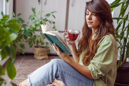 Téléchargez les photos : Belle jeune femme assise sur son balcon à boire du thé et à lire un livre tout en se relaxant à la maison - en image libre de droit