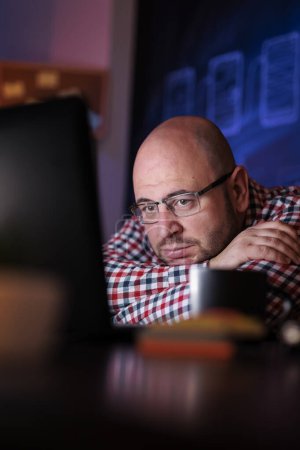 Foto de Hombre sentado en su escritorio en la oficina del hogar, cansado mientras trabaja hasta tarde en el desarrollo de nuevas aplicaciones, apoyado en el escritorio y mirando la pantalla del ordenador portátil - Imagen libre de derechos