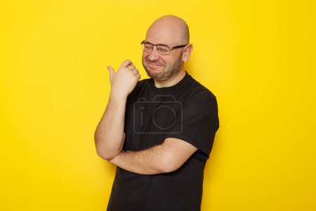 Photo for Portrait of cheerful funny mid 30s bald overweight man wearing black T-shirt and glasses making faces isolated on yellow colored background - Royalty Free Image