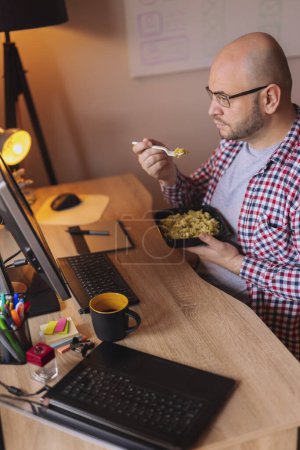 Téléchargez les photos : Homme assis à son bureau dans un bureau, prenant une pause déjeuner, mangeant des pâtes à emporter et buvant du café - en image libre de droit