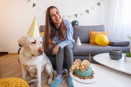 Téléchargez les photos : Femme propriétaire d'animal de compagnie s'amuser à faire une fête d'anniversaire pour son chien à la maison, chien portant un chapeau de fête et soufflant bougie sur le gâteau d'anniversaire - en image libre de droit