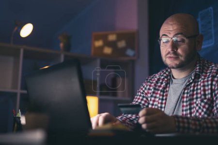 Photo for Man sitting at his desk late at night, using laptop computer and paying bills online, inserting credit card number while making payments - Royalty Free Image
